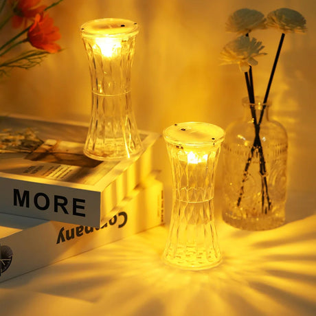 Two glass candle holders with lit candles on a surface, next to a book and a vase with flowers.